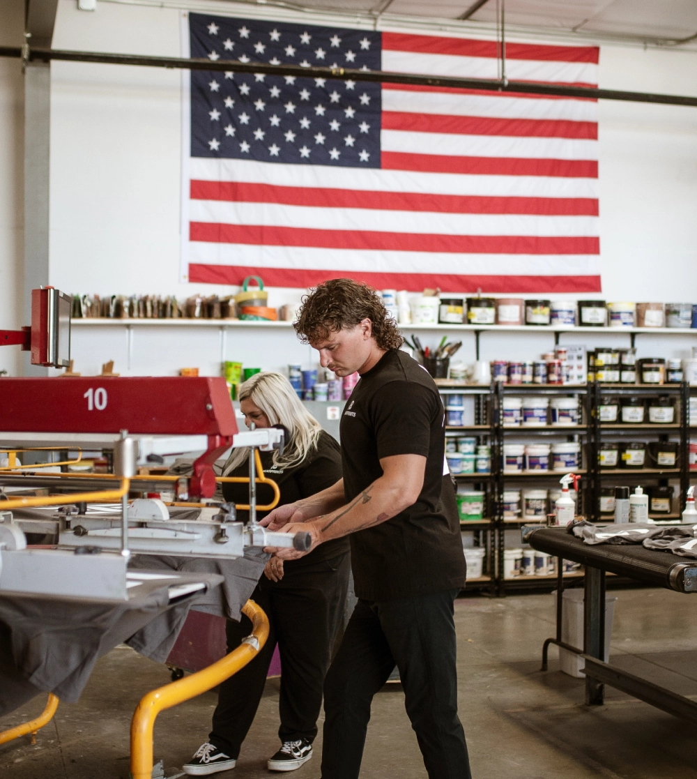 Two people working in a shop with an american flag on the wall.